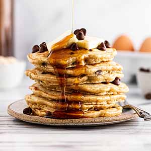 pouring maple syrup on a stack of oatmeal chocolate chip pancakes.