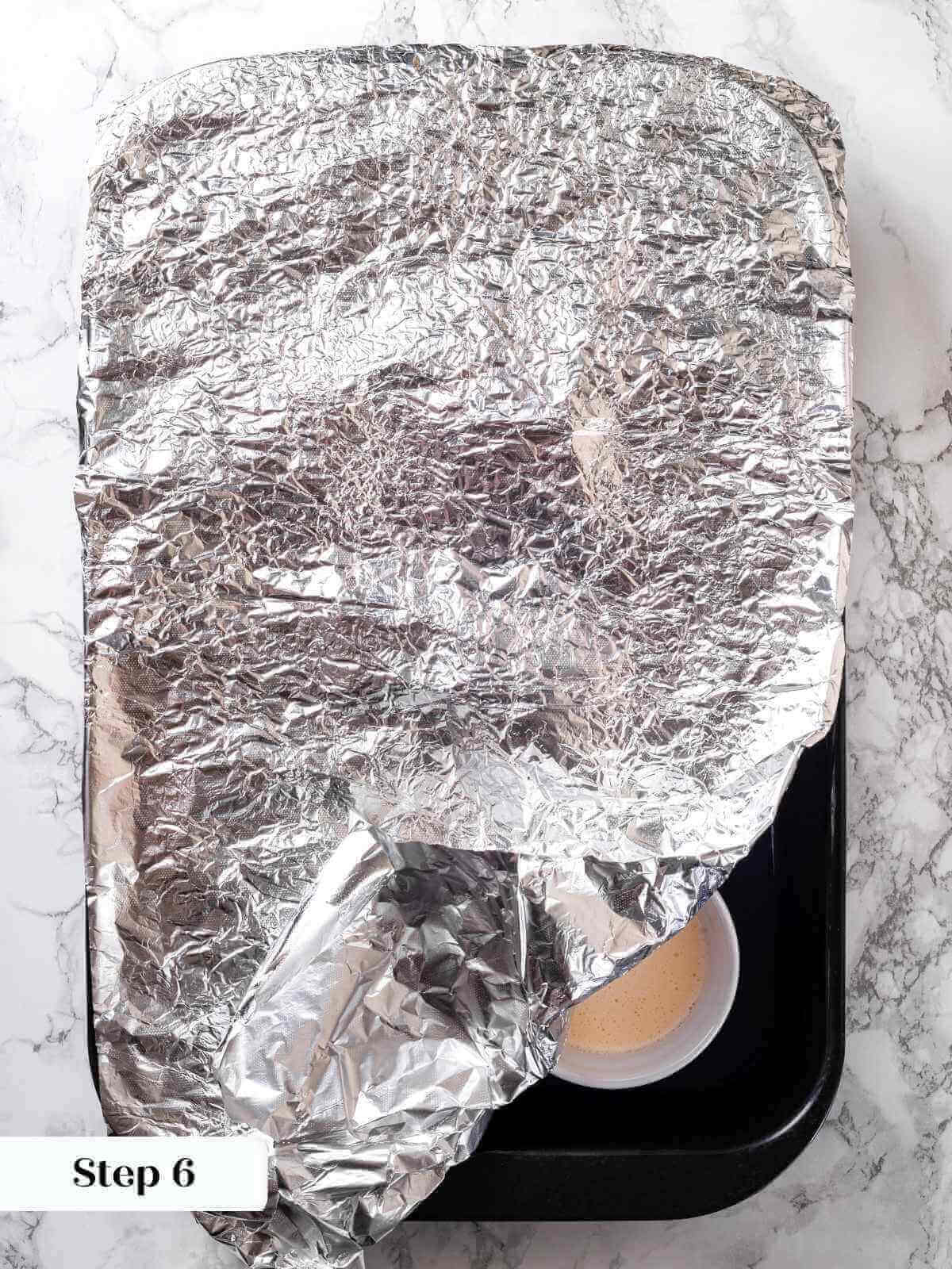 A baking dish covered with foil, holding creme brulee ramekins for gentle baking.
