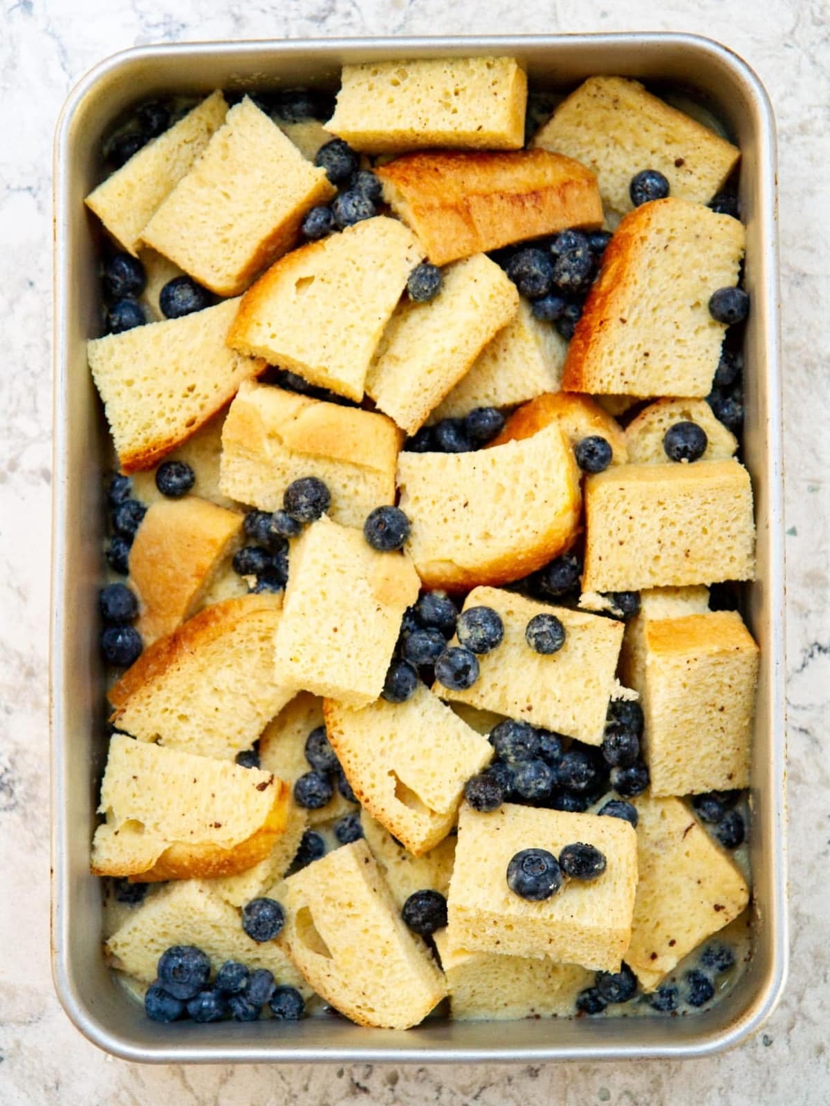 Unbaked casserole with brioche cubes, blueberries, and custard mixture evenly poured across the dish.