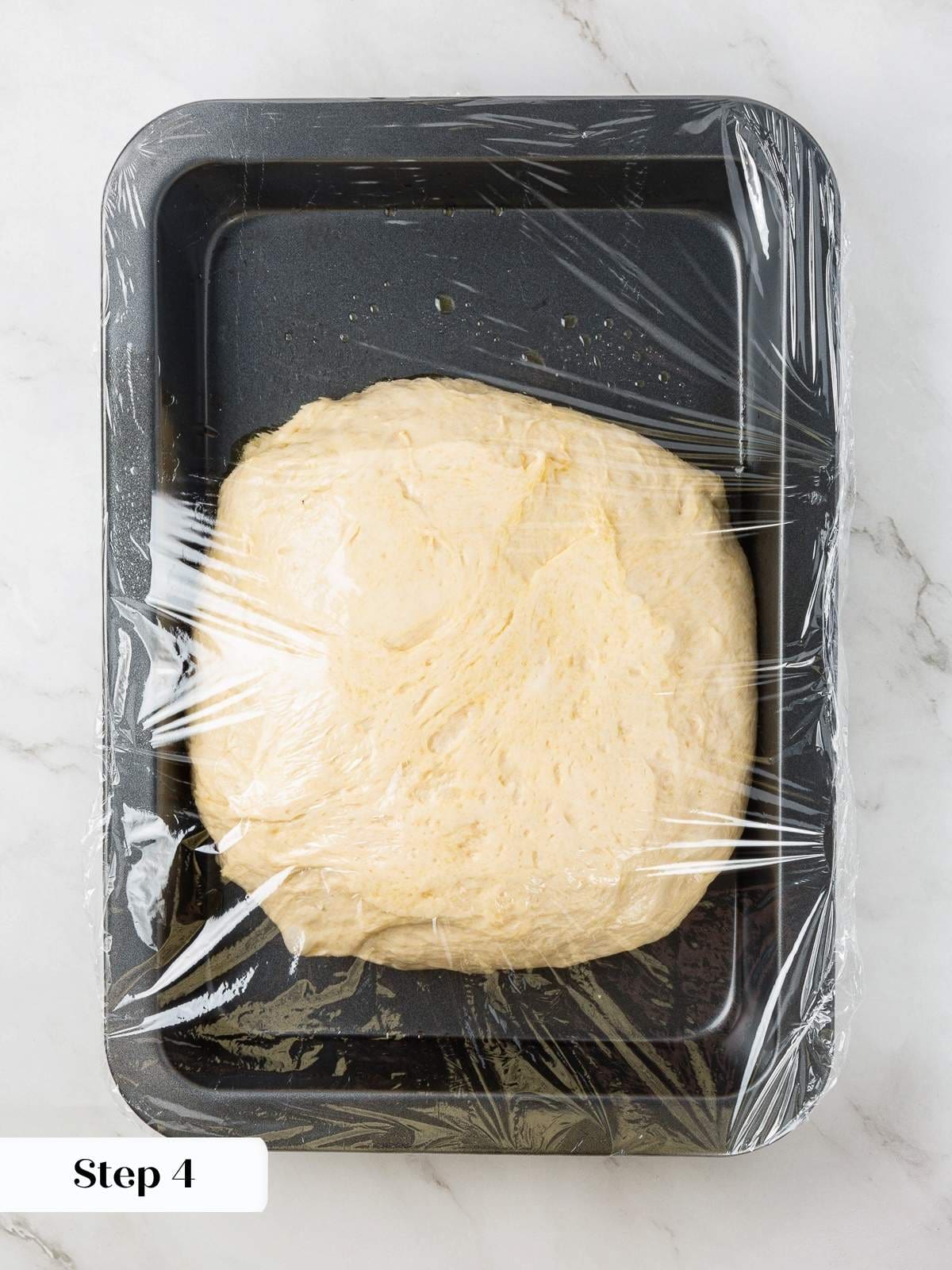 Focaccia dough covered in plastic wrap and ready to rise in a warm kitchen.