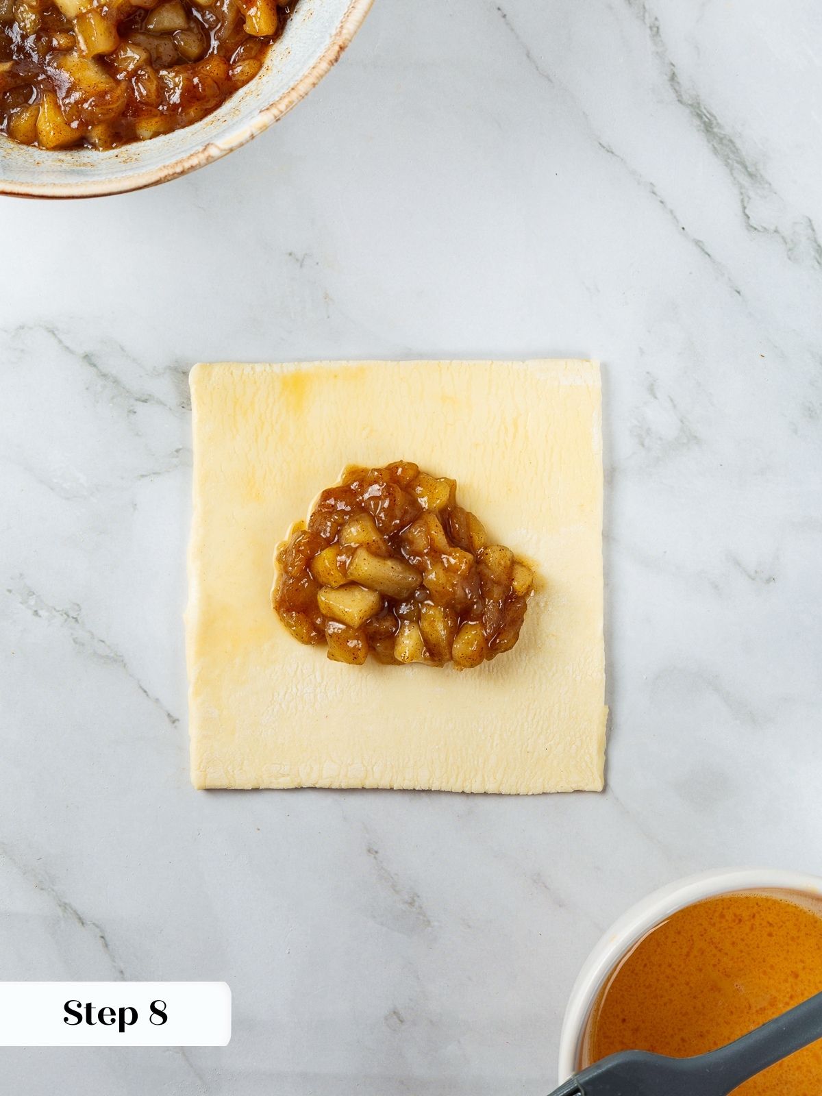 Spoonful of spiced apple filling being placed onto puff pastry square before folding.