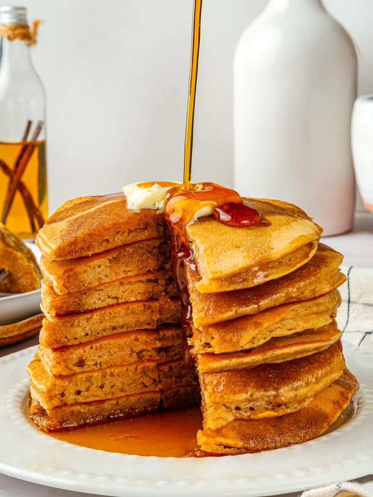 Maple syrup being poured generously over a stack of fluffy pumpkin pancakes with butter.