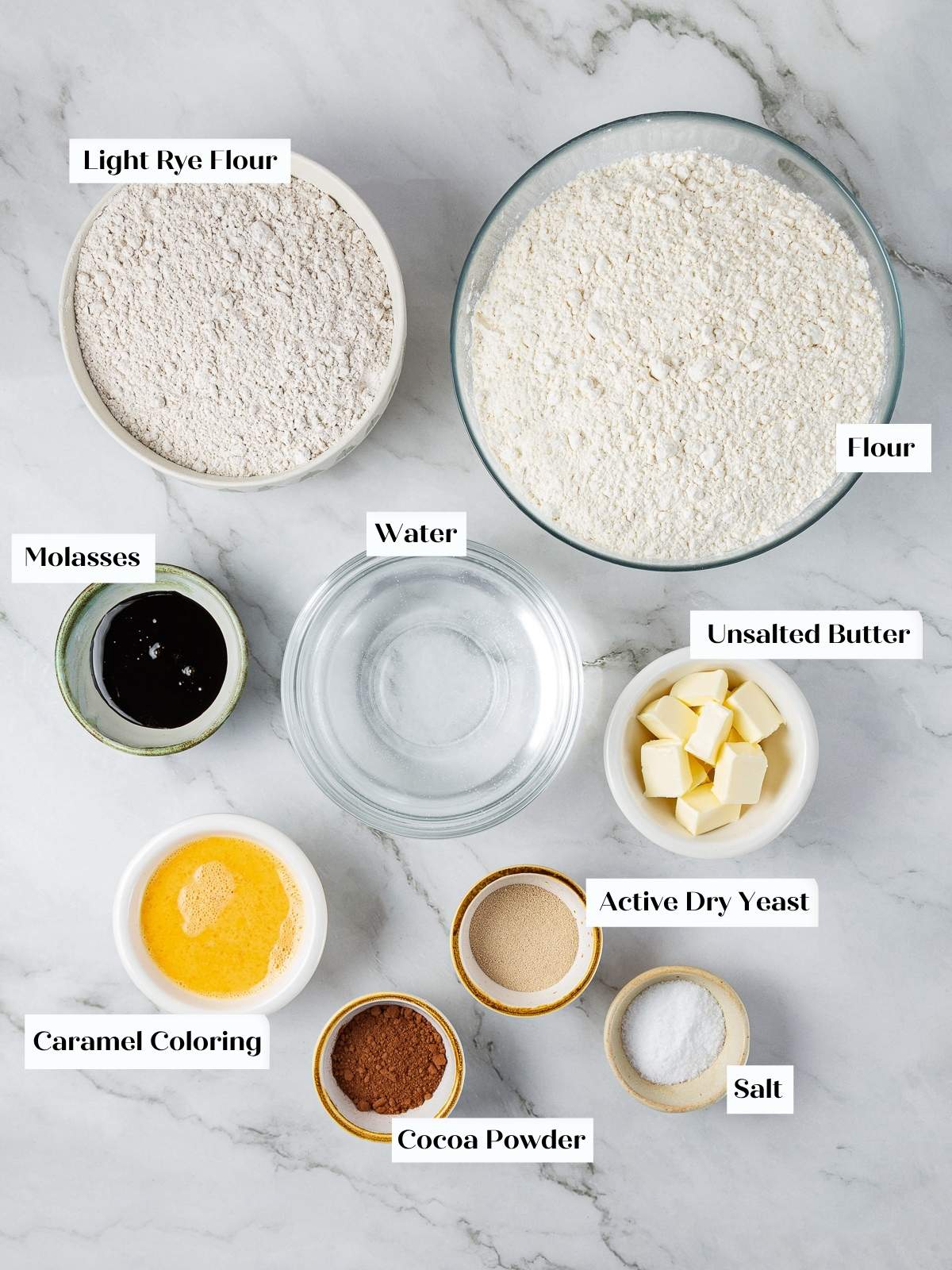 Ingredients for marble rye bread arranged on a kitchen counter including flours, molasses, butter, and yeast.