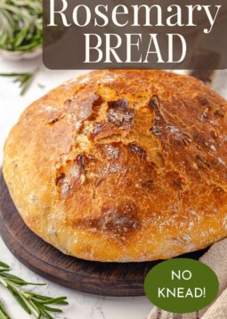 Round loaf of rosemary bread on a cutting board surrounded by fresh rosemary.