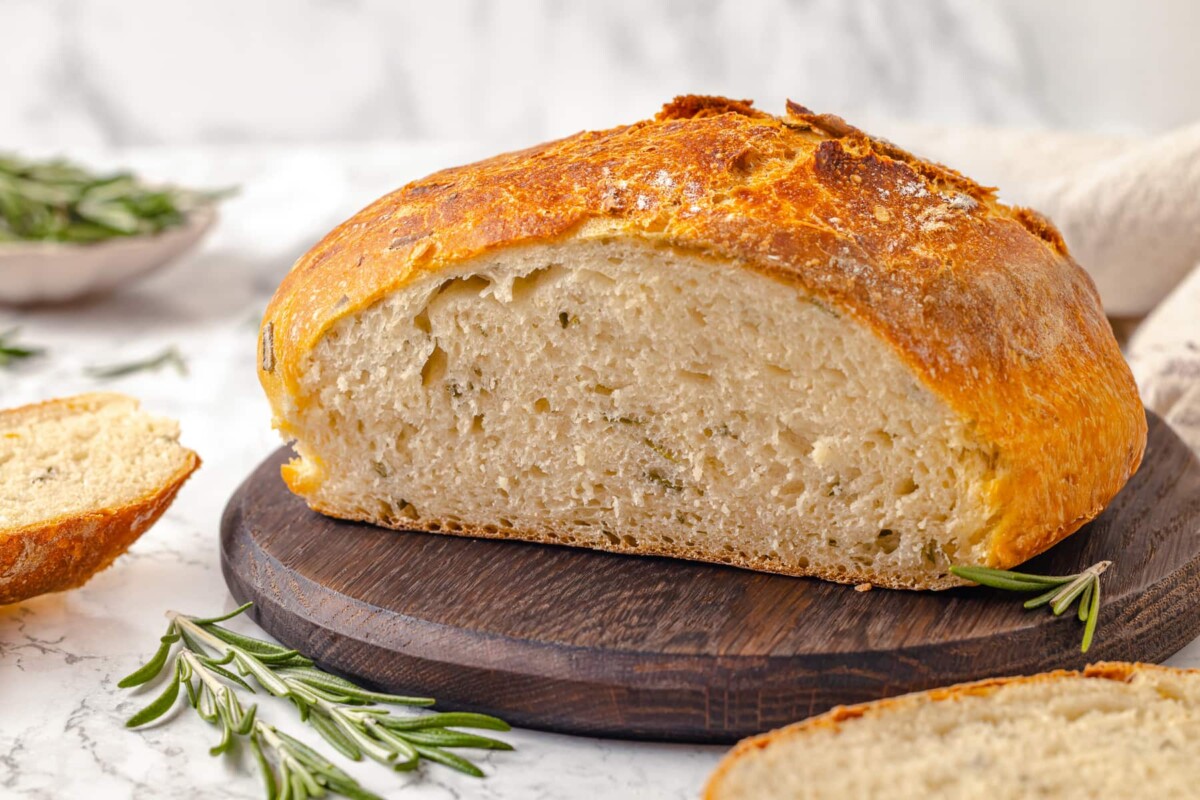 Loaf of rosemary bread resting on a wooden board beside sprigs of fresh herbs.