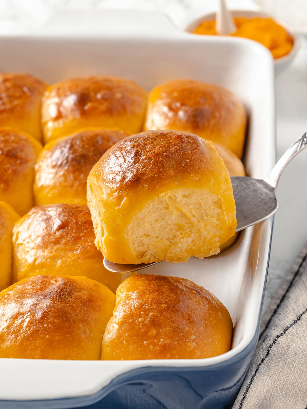 Sweet potato rolls being lifted from a baking dish with a spatula beside melted butter.