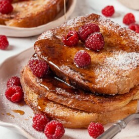 Pouring maple syrup over a stack of warm brioche French toast on a white breakfast plate.