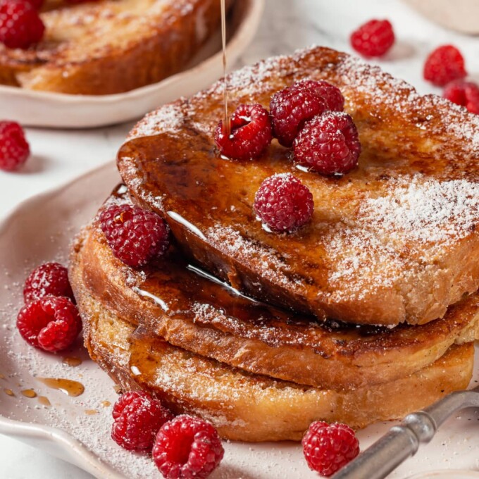 Pouring maple syrup over a stack of warm brioche French toast on a white breakfast plate.