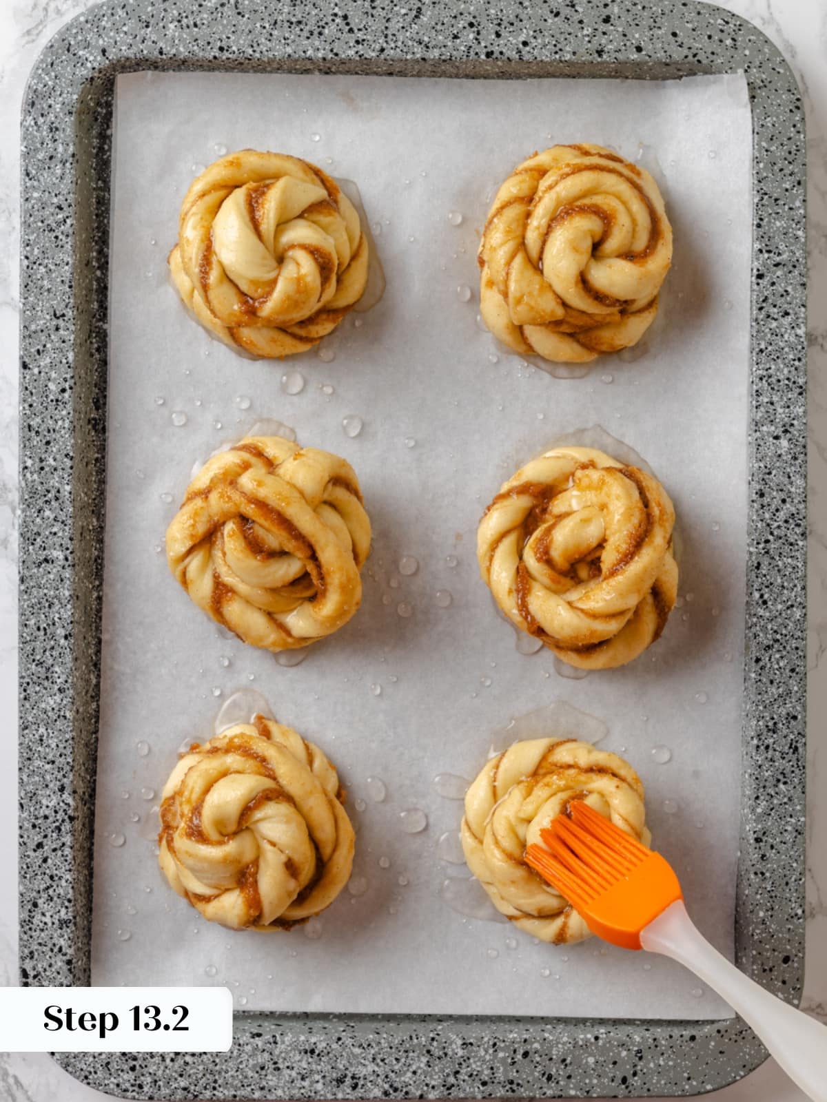 Brushing unbaked buns with sugar syrup for a sweet caramelized finish.
