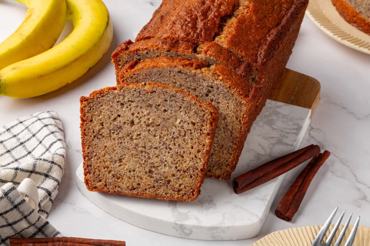 Sliced sourdough banana bread loaf displayed with cinnamon sticks beside board.