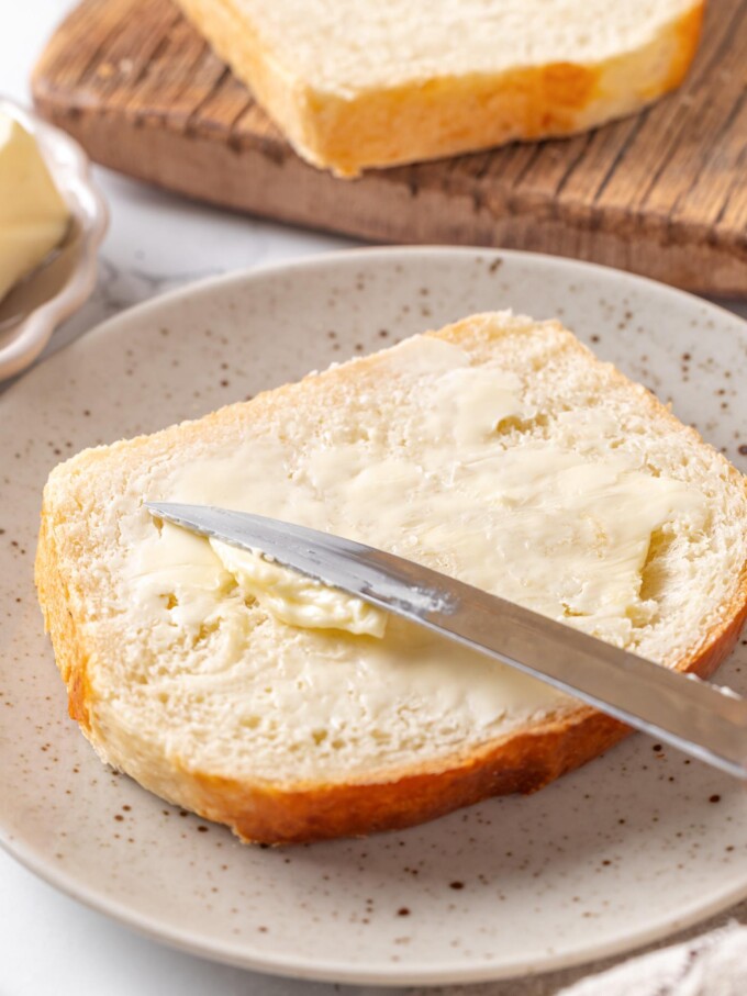 Slice of buttermilk bread on plate being spread with butter using metal table knife