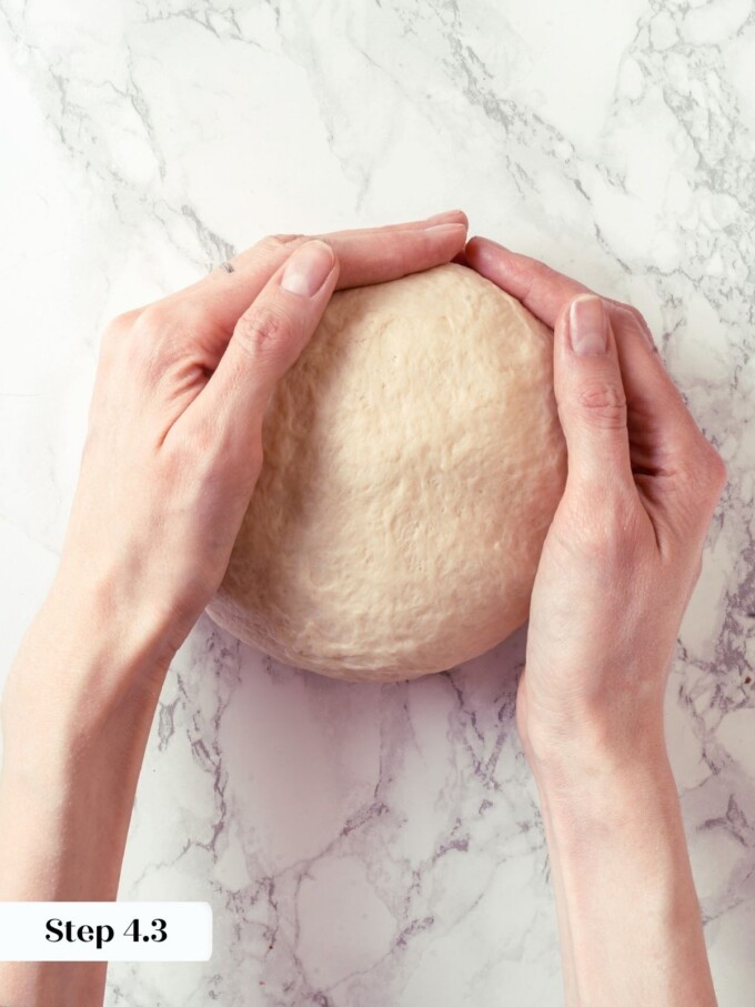 Two hands rounding buttermilk dough into a tight ball on white marble surface.