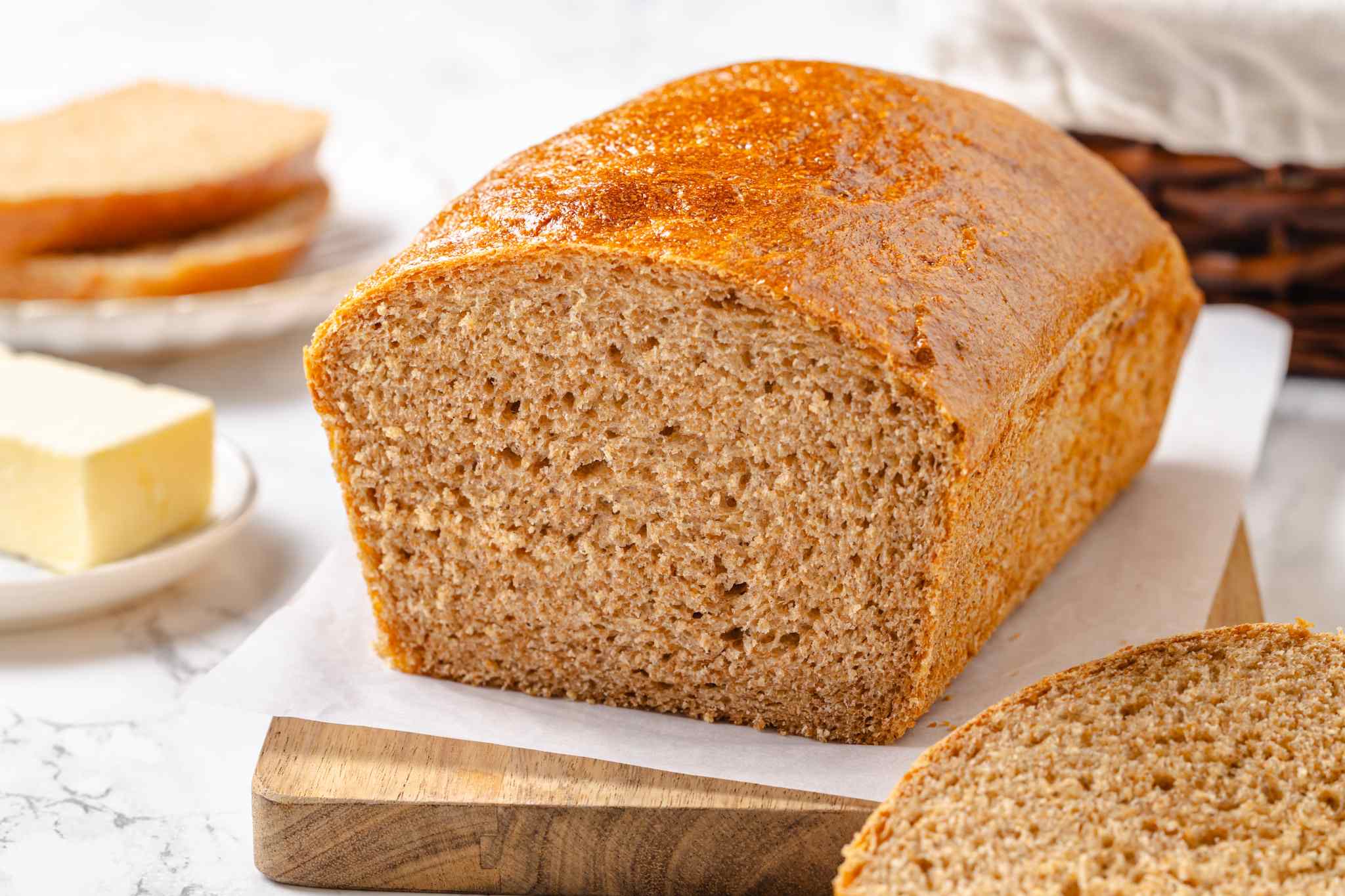 Fluffy whole wheat loaf sliced with butter and bread staged across white surface.