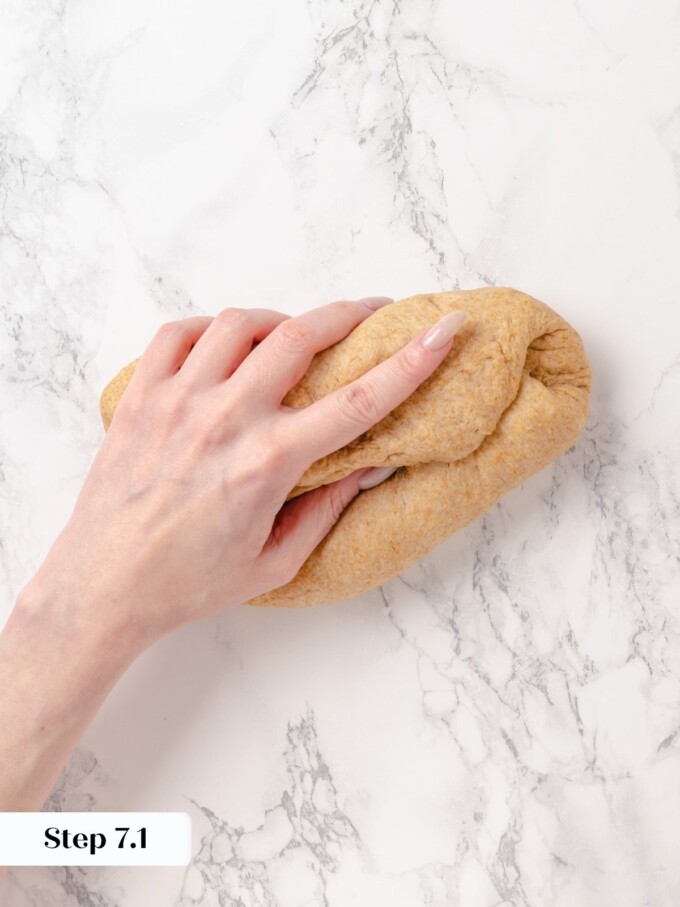 Hand pressing folded whole wheat bread dough flat on white marble counter.