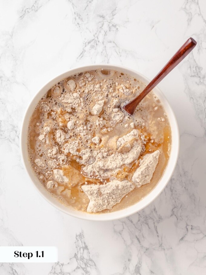 Whole wheat flour scattered over yeast water in white bowl with wooden spoon.