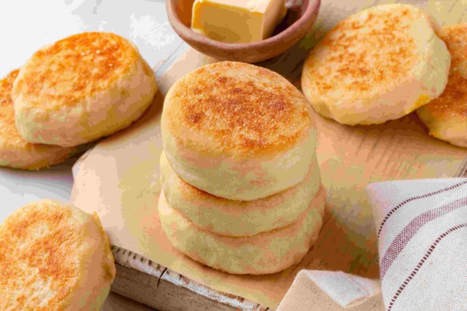 Stack of sourdough English muffins surrounded by additional muffins, butter dish, and a striped linen towel.