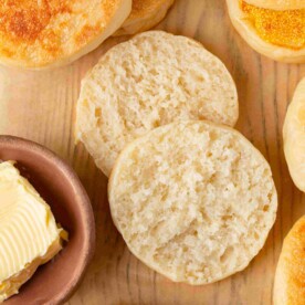 Sourdough English muffin halves split open on wood board showing tender crumb with butter dish nearby.