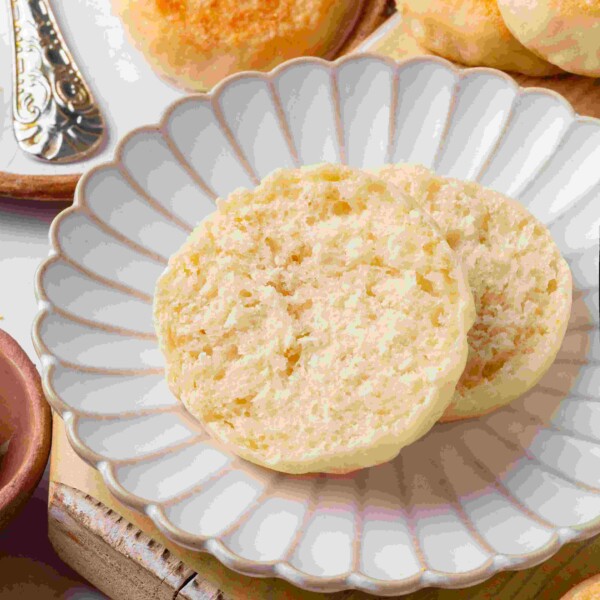 Split sourdough English muffin halves on a white scalloped plate surrounded by whole muffins and butter.