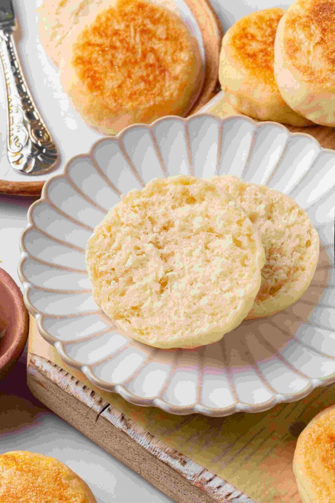 Split sourdough English muffin halves on a white scalloped plate surrounded by whole muffins and butter.