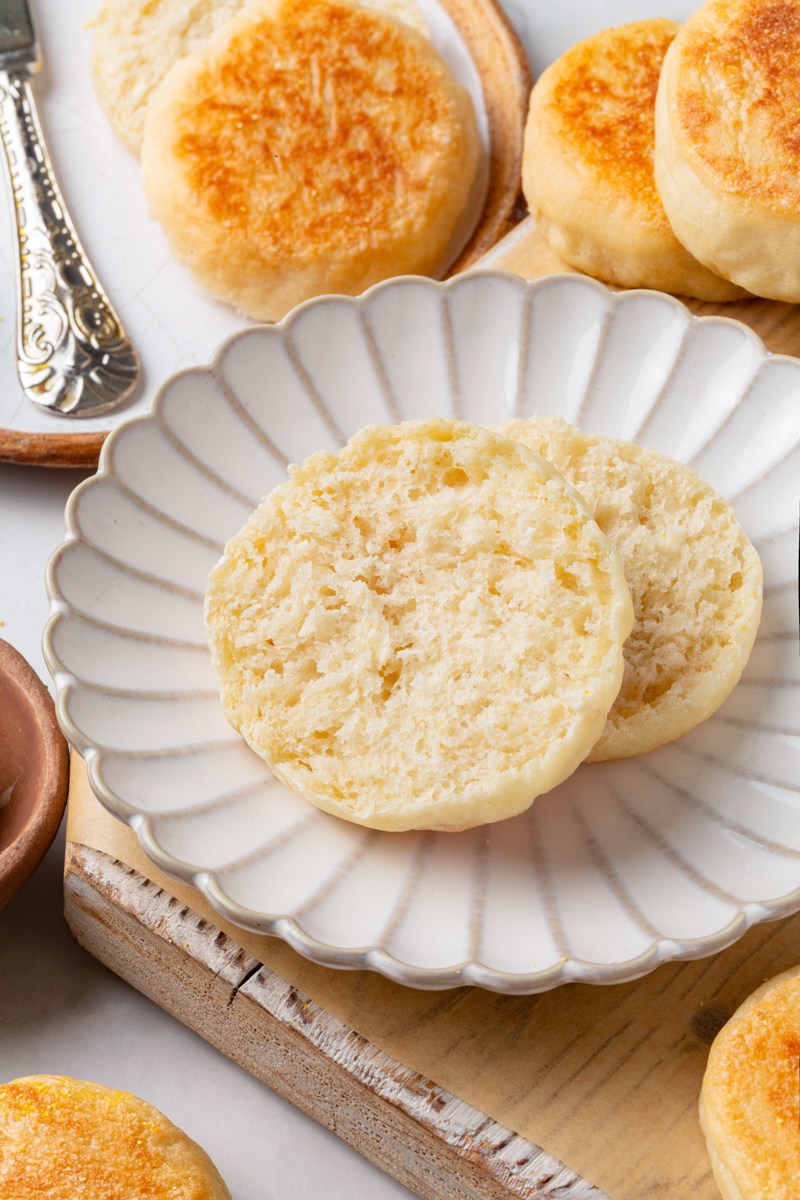 Split sourdough English muffin halves on a white scalloped plate surrounded by whole muffins and butter.
