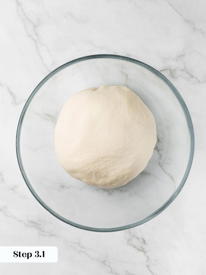 Smooth dough ball resting in a glass bowl on marble before the first bulk proof begins.