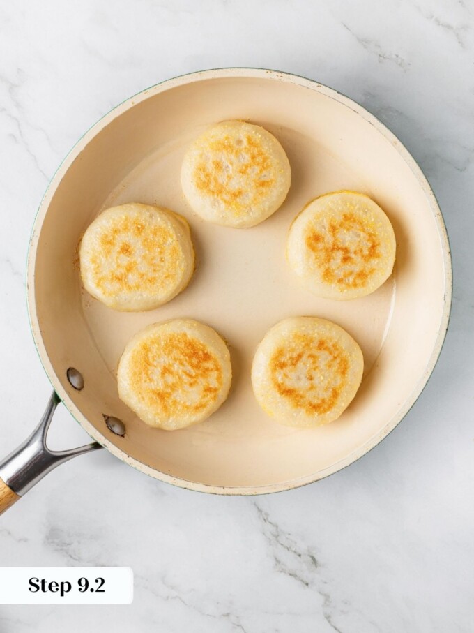 Five sourdough English muffins flipped in skillet showing golden brown cornmeal-crusted bottoms after pan frying.