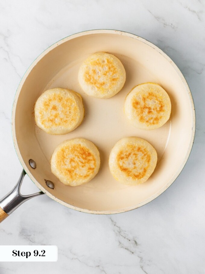 Five sourdough English muffins flipped in skillet showing golden brown cornmeal-crusted bottoms after pan frying.