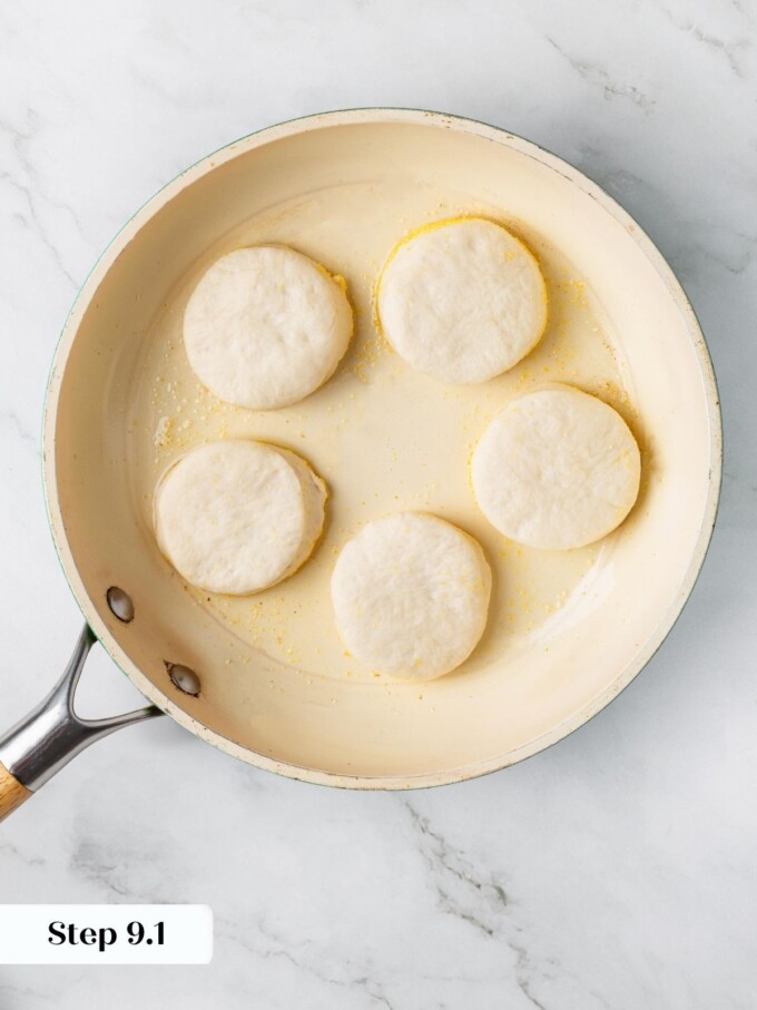 Five sourdough English muffin rounds cooking in a cream colored skillet with white sides facing up.