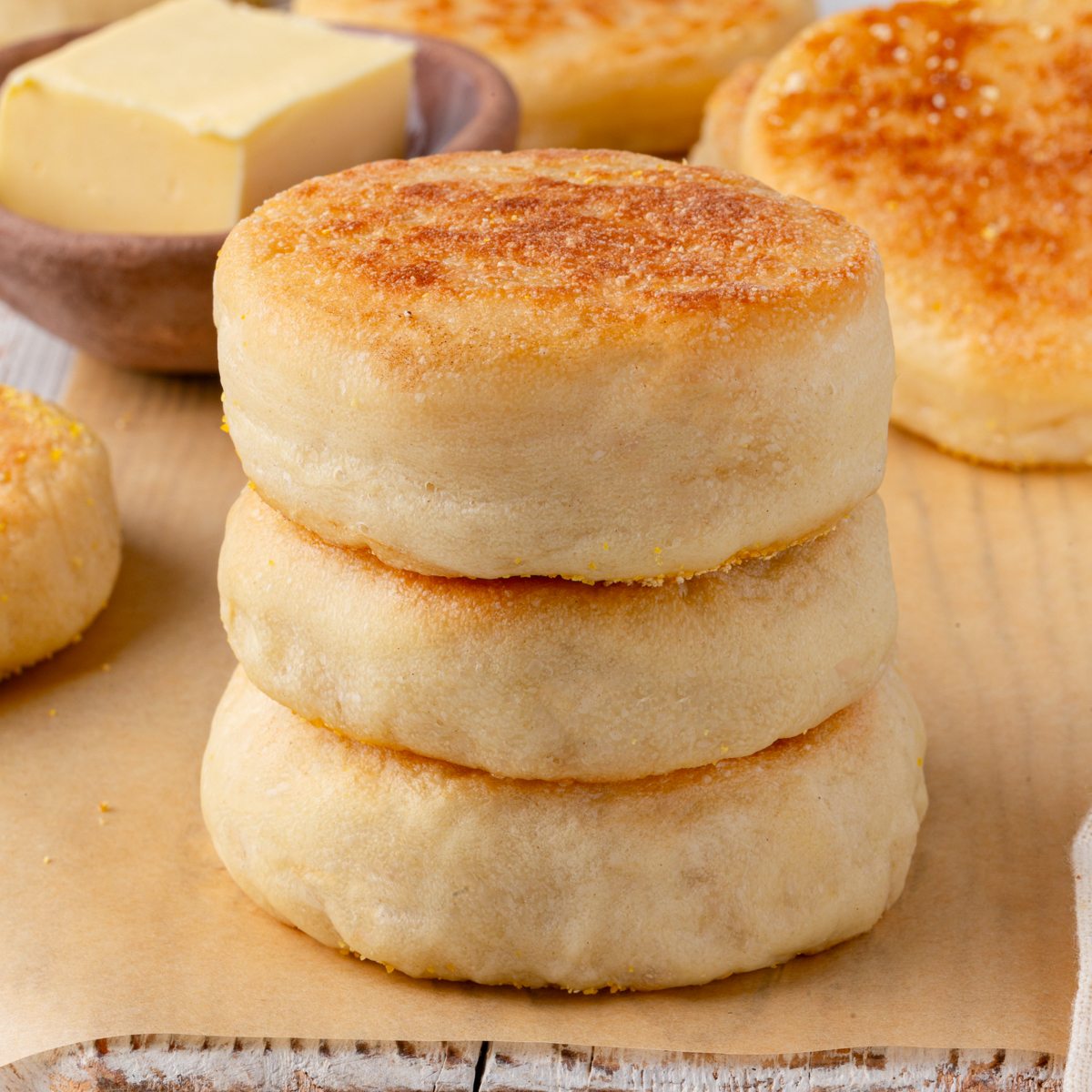 Three sourdough English muffins stacked on parchment paper with a butter dish in the background.