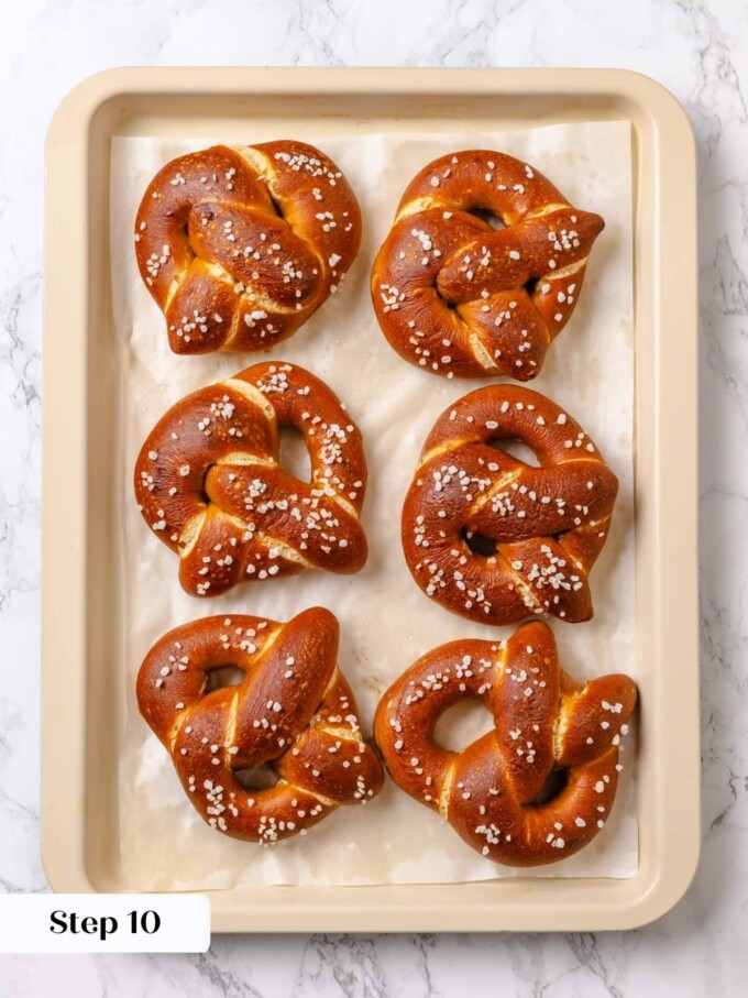 Six deeply browned baked pretzels with coarse salt arranged on parchment inside a cream sheet pan.