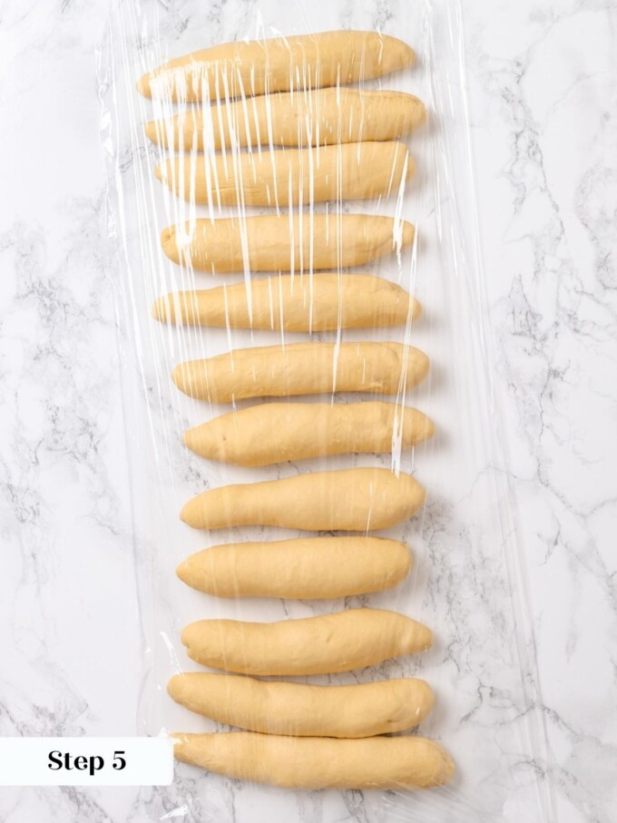 Twelve preshaped dough logs covered loosely in plastic wrap to bench rest before final rolling.