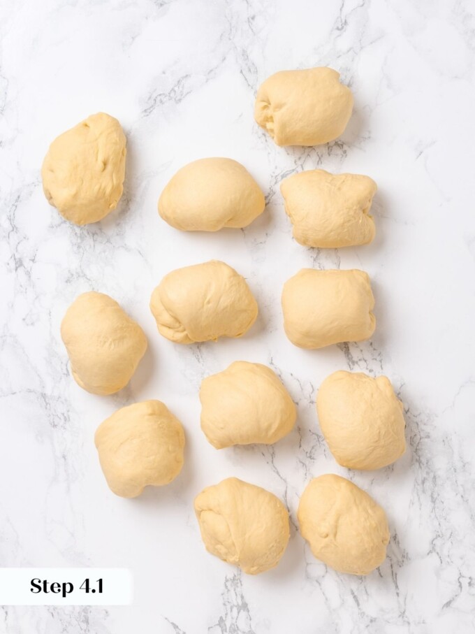 Twelve equal portions of pale pretzel dough scattered loosely across a white marble work surface.