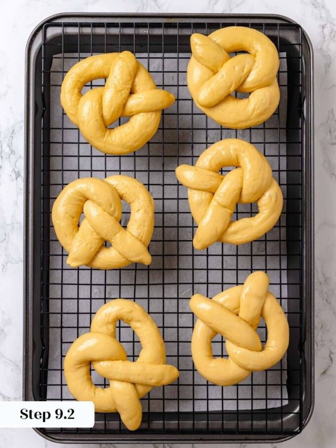 Six lye-dipped pretzels resting on a black wire rack to drip before transferring to baking parchment.