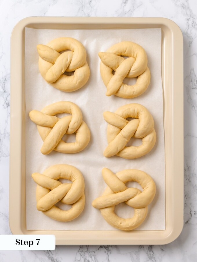 Six shaped pretzels softly risen after final proofing on parchment inside a cream rimmed sheet pan.