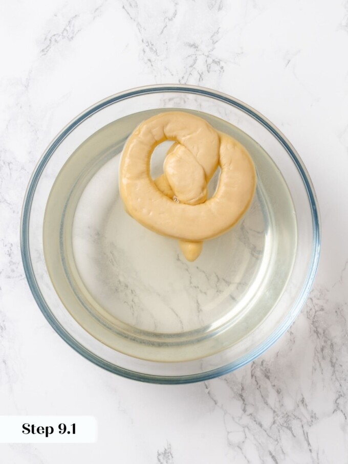 A single shaped pretzel submerged face-down in a clear glass bowl filled with lye solution.