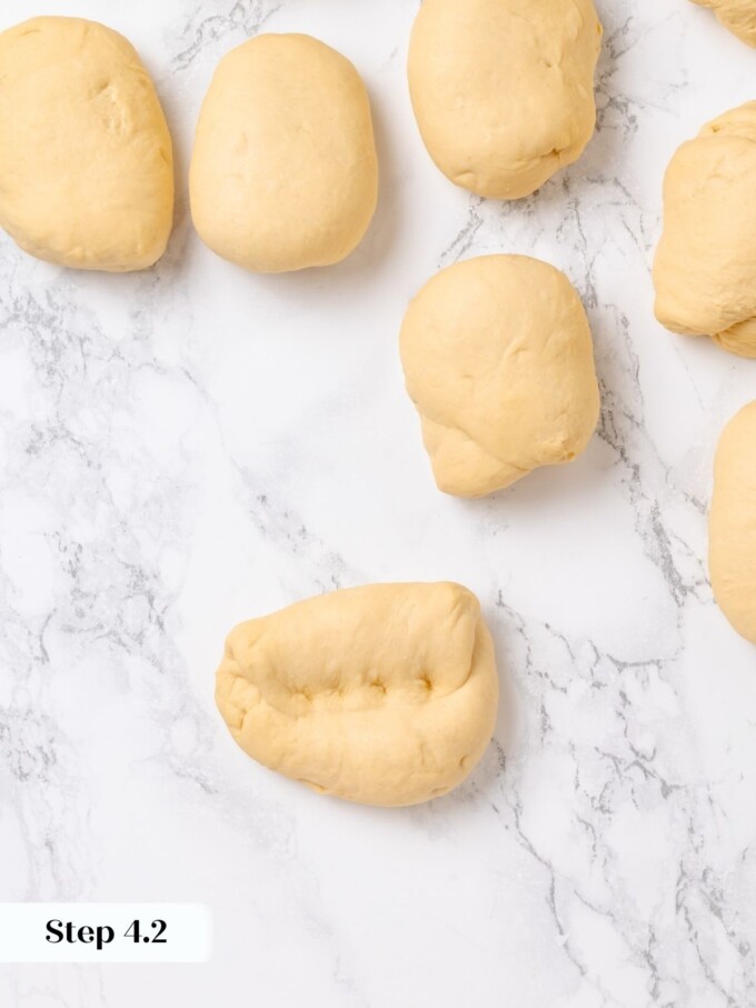 Several preshaped dough pieces with one freshly folded showing the seam on a marble countertop.