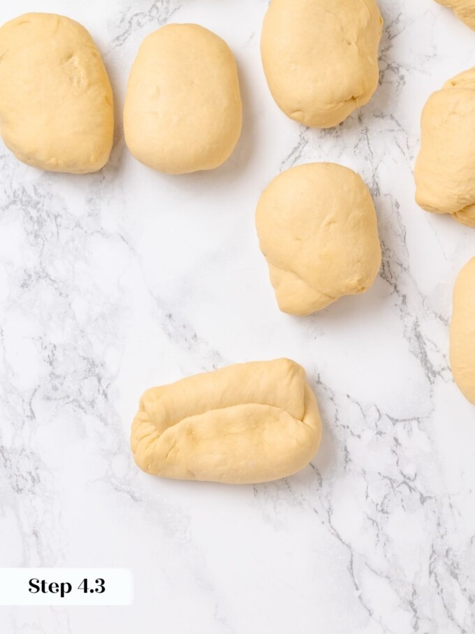 Preshaped dough pieces with one rolled into a stubby rope on a marble work surface.