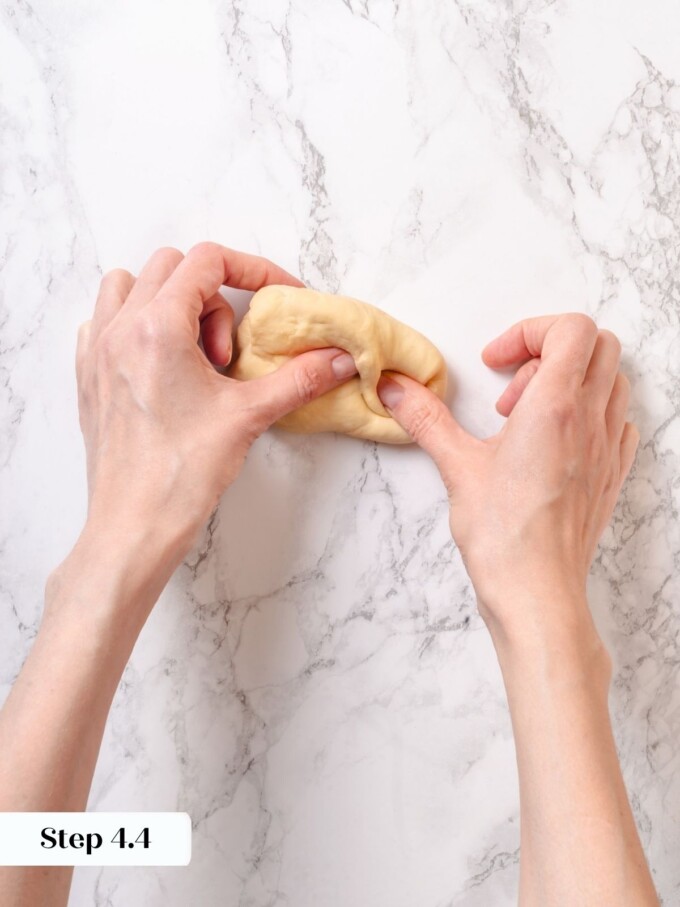 Hands working a thumb-fold into a small piece of pale pretzel dough on white marble.