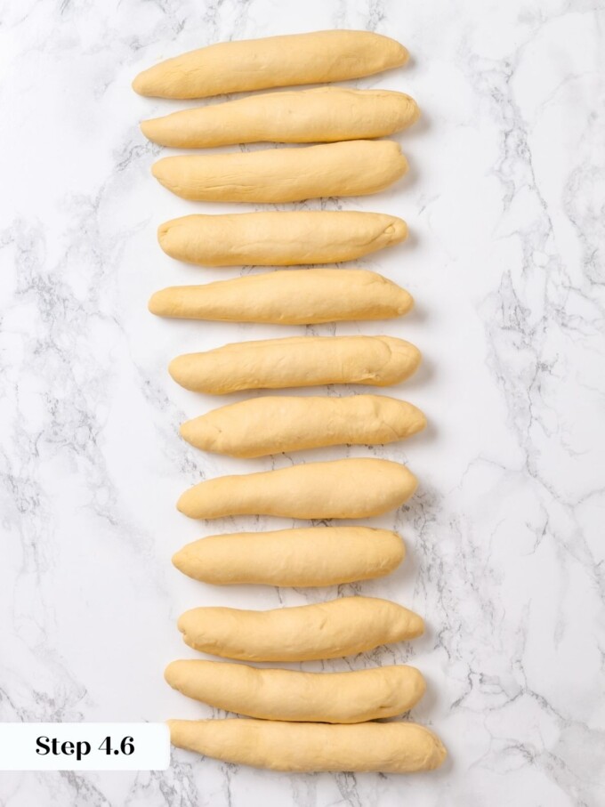 Twelve preshaped pretzel dough logs lined up in a single vertical row on white marble.