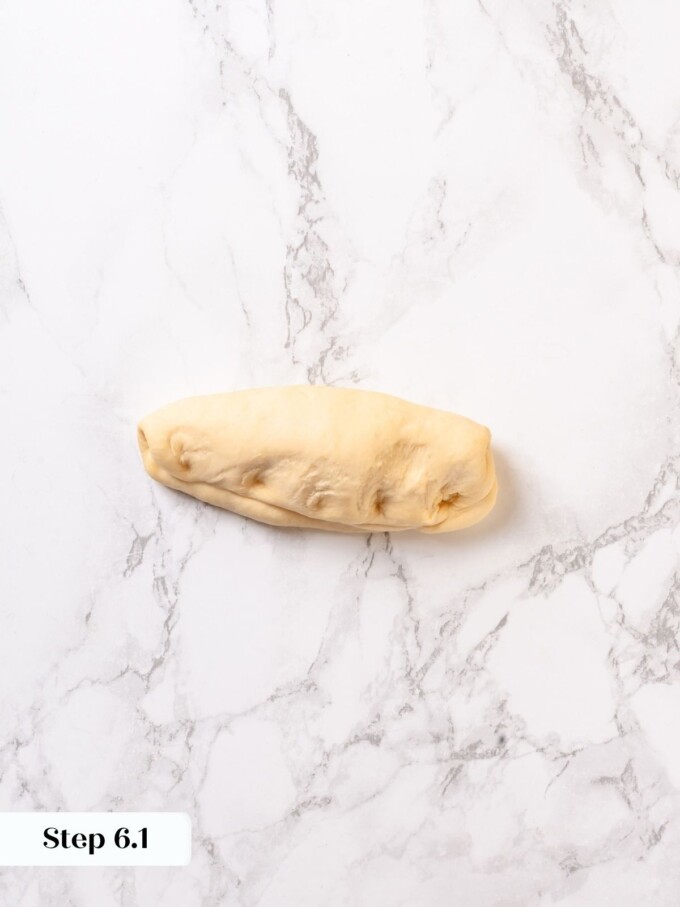 A single pre-shaped pretzel dough log resting on white marble before rolling into a rope.