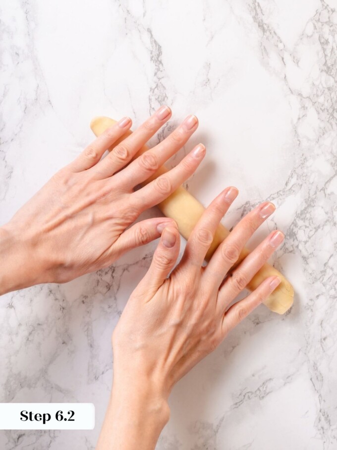 Hands rolling a piece of pretzel dough into a long even rope on a marble surface.