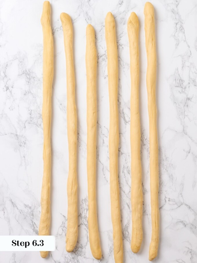 Six long pretzel dough ropes laid parallel and ready for shaping on a marble work surface.