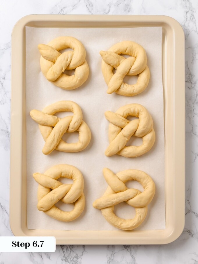Six raw shaped pretzels resting in two rows on parchment paper inside a cream sheet pan.