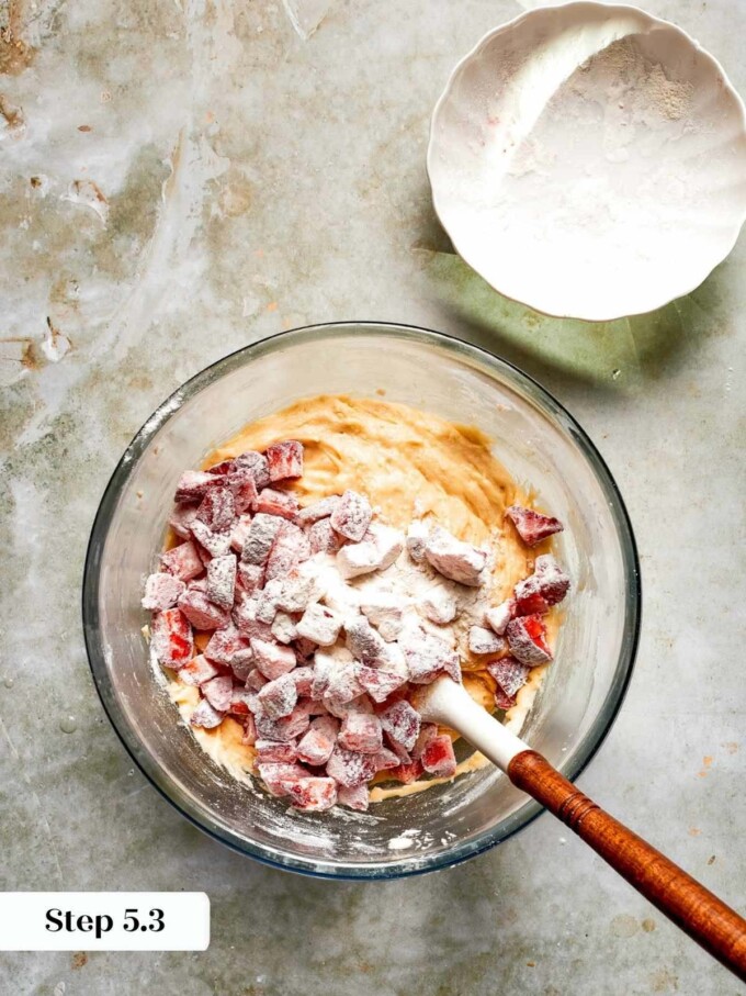 Coated diced strawberries added to smooth batter in a glass bowl, ready to be gently incorporated.