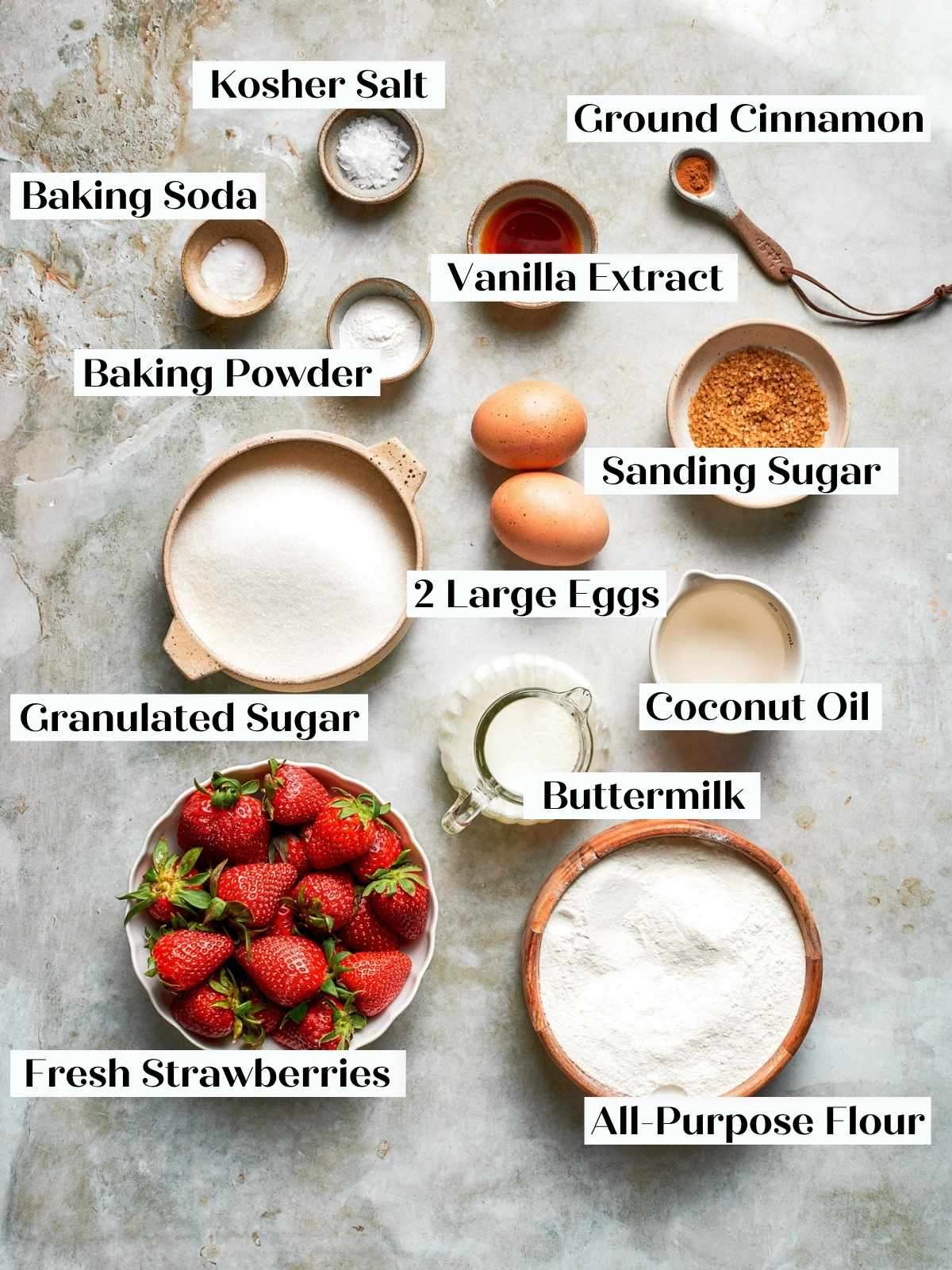 Ingredients for strawberry bread arranged on a stone surface, including flour, sugar, eggs, and fresh strawberries.
