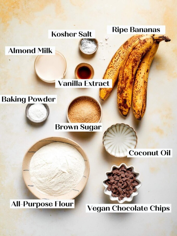 Vegan banana bread ingredients on a beige surface: flour, ripe bananas, brown sugar, chocolate, plant milk.