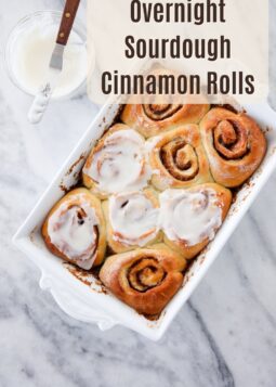 Overhead view of sourdough cinnamon rolls in a white baking dish, half frosted, with a bowl of cream cheese frosting